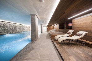 A modern indoor pool area with a stone wall, wooden deck, and lounge chairs. - Hotel Touring Livigno (Livigno)