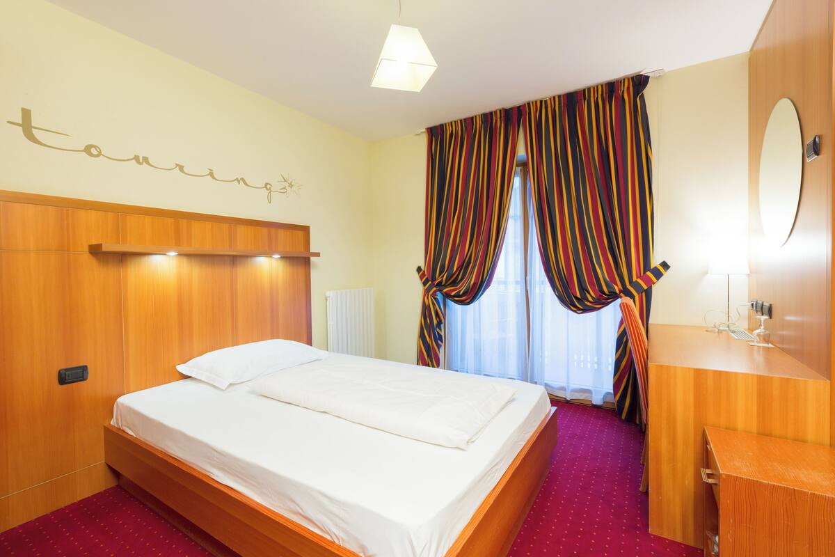 A hotel room with a wooden bed, a bedside table, a mirror, and a window with striped curtains.