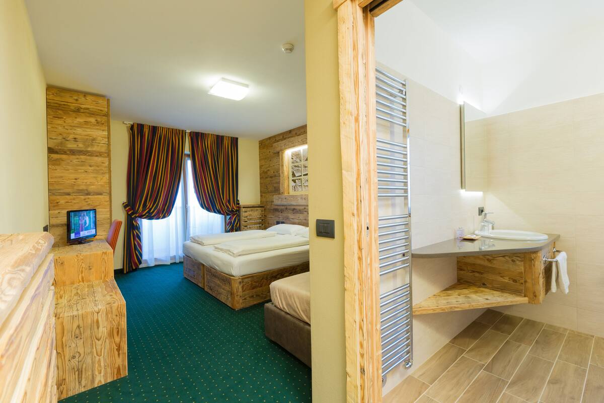 A hotel room with a wooden bed, a built-in TV, a small desk, a bathroom with a wooden vanity and a towel rack.