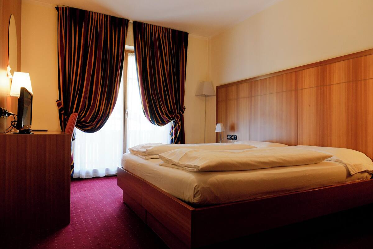 A hotel room with a wooden headboard, a bed with white linens, a desk with a lamp, and curtains.