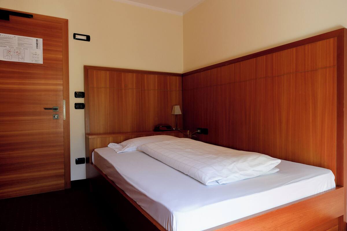 A hotel room with a wooden headboard, a single bed, a nightstand with a lamp, and a door with a notice.