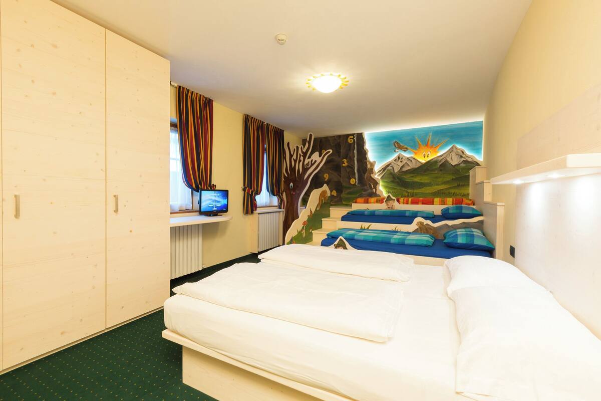 A room with a bed, a wardrobe, a window with curtains, a television, and a mural on the wall.