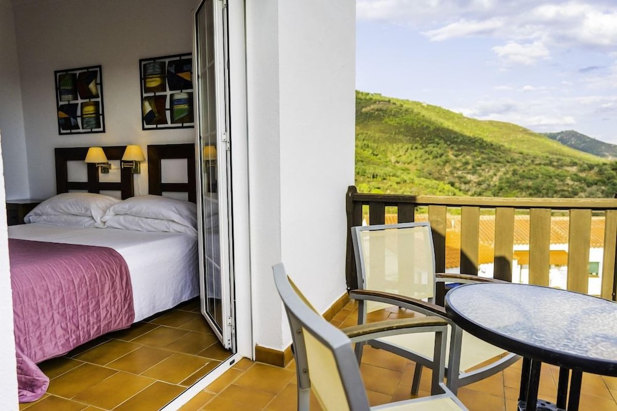 A hotel room with a bed, a table, chairs, and a balcony with a view of green hills.