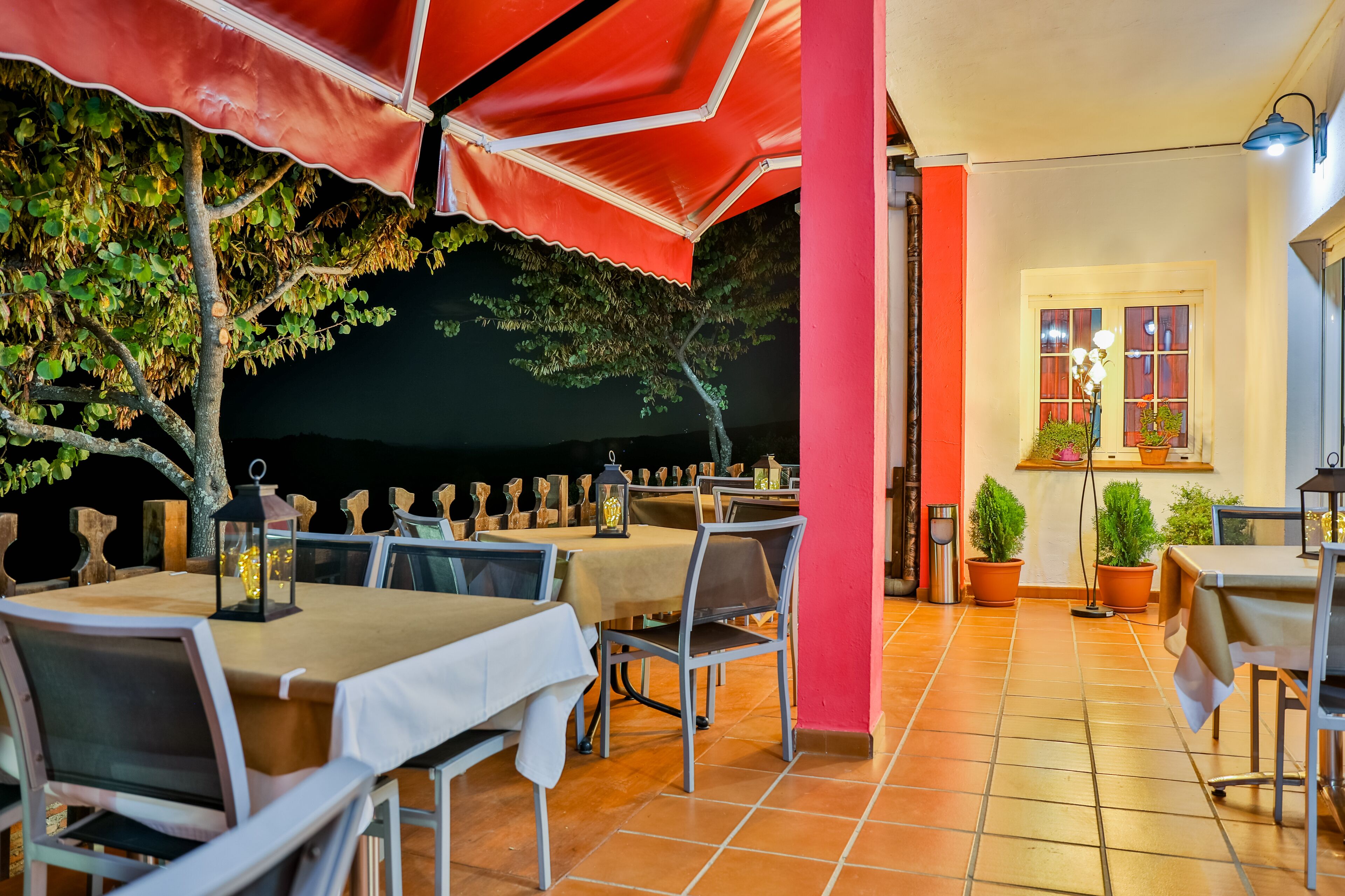Un área de comedor al aire libre con mesas y sillas, sombrillas rojas y plantas en macetas.