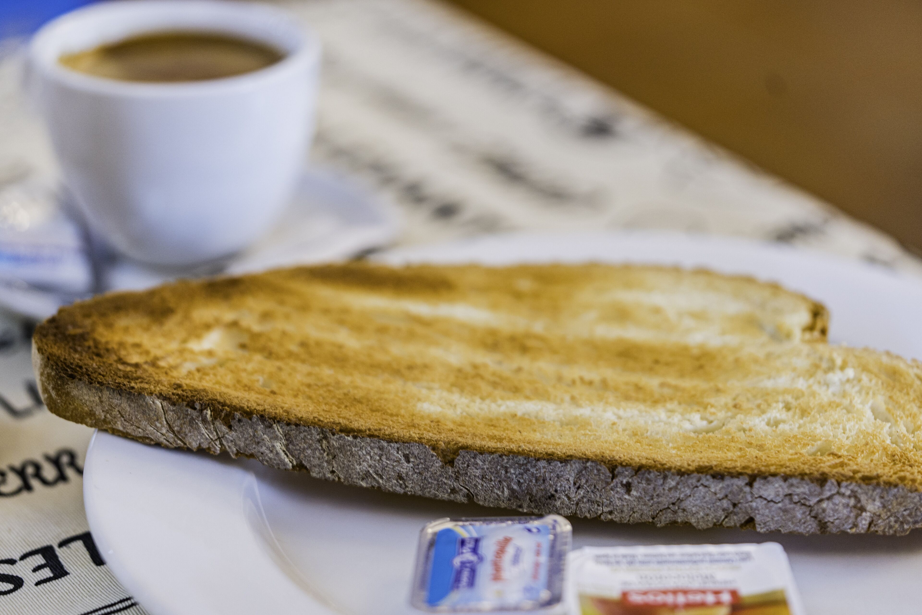Una rebanada de pan tostado en un plato con un recipiente pequeño de mantequilla y una taza de café.