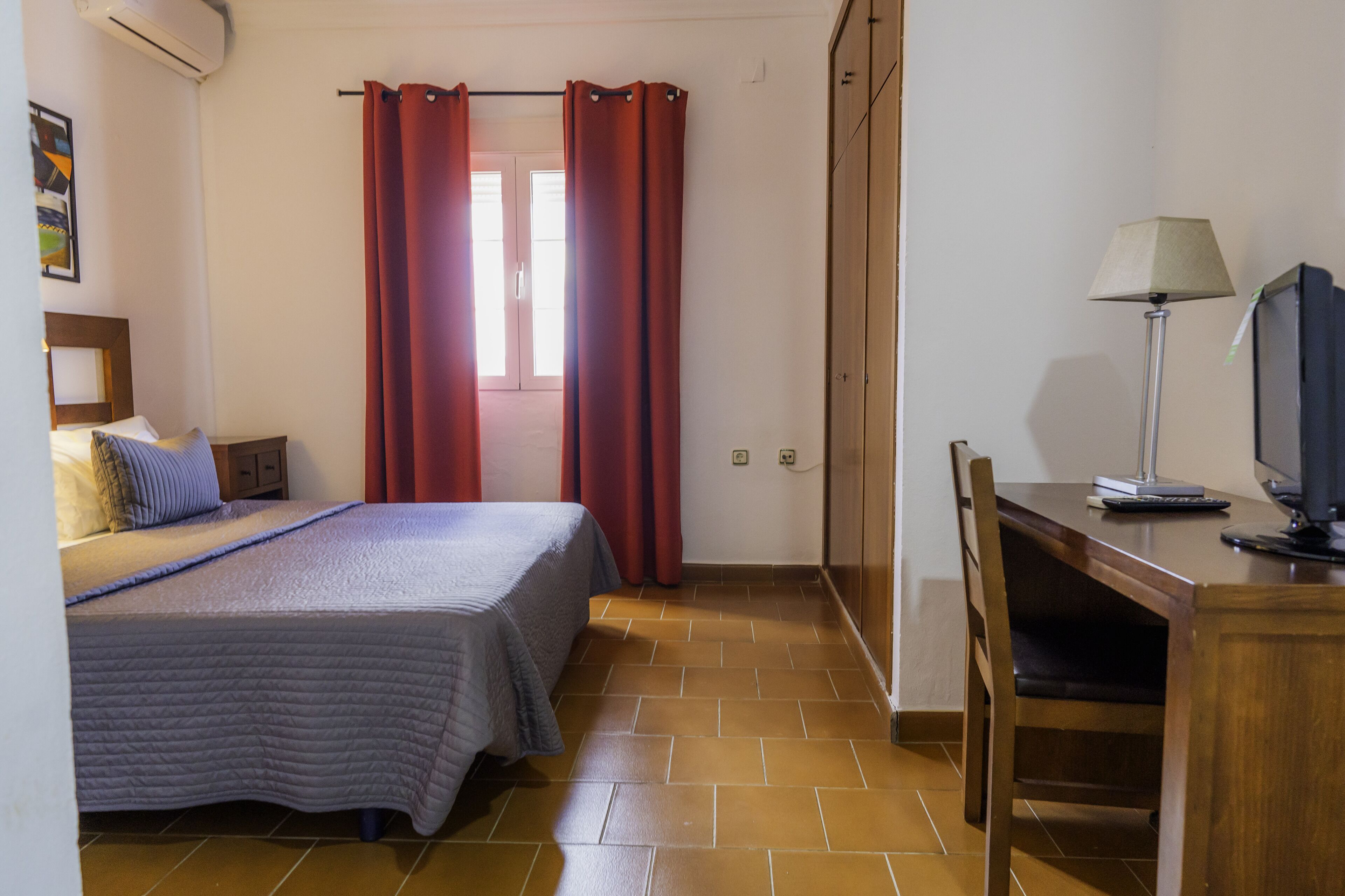 A hotel room with a bed, a desk with a lamp, a TV, and a window with red curtains.