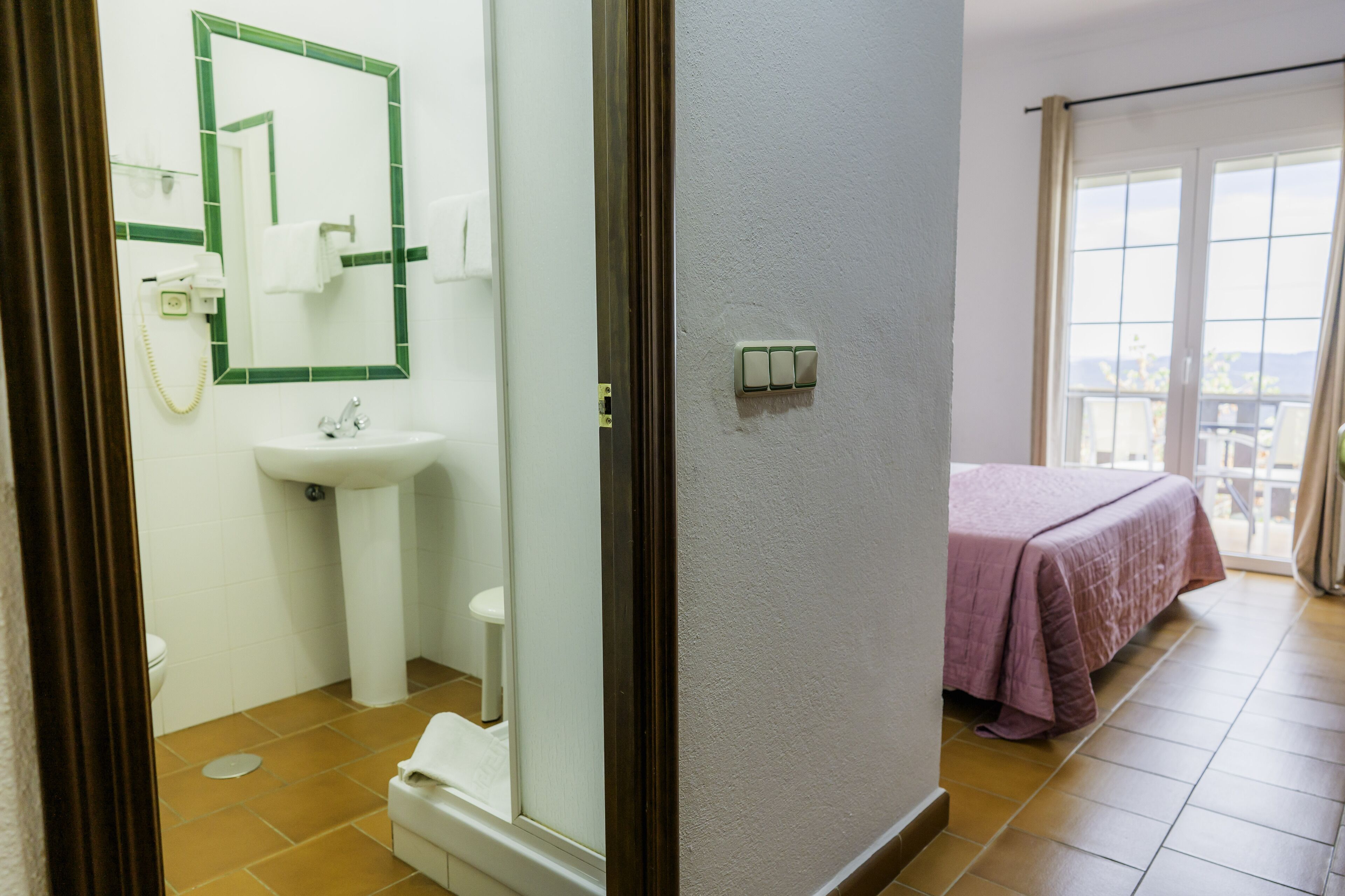 A bathroom with a sink, mirror, and toilet. There is a bed with a pink bedspread in the adjacent room.