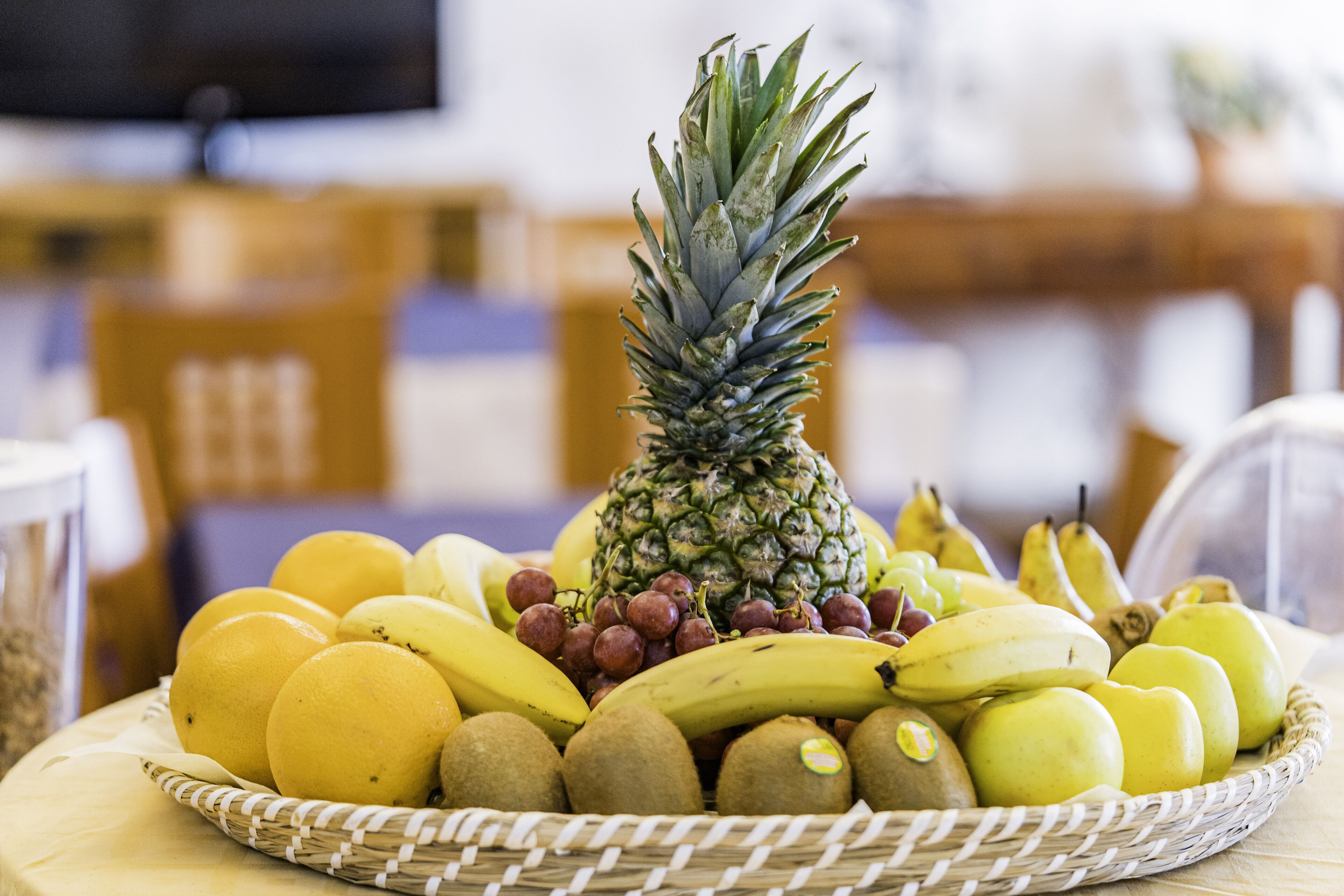 A fruit basket with a pineapple, bananas, kiwis, and oranges on a table.
