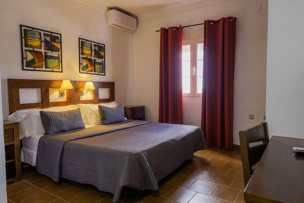 A hotel room with a bed, two bedside lamps, a window with red curtains, and two abstract paintings on the wall.
