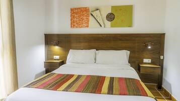 Une chambre d’hôtel avec une tête de lit en bois, un lit recouvert de draps blancs, une couverture rayée aux couleurs vives, deux tables de chevet avec des lampes et des œuvres d’art accrochées au mur.