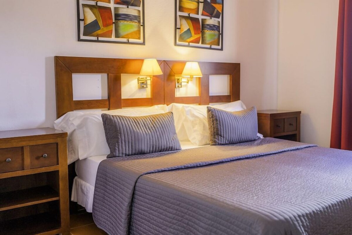 A hotel room with a wooden headboard, two bedside lamps, a bed with a quilted cover, and a nightstand with a drawer.