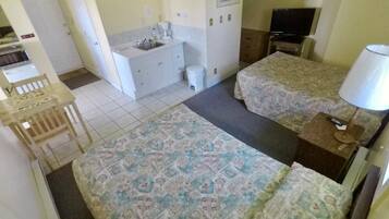 A small, compact room with two beds, a desk, a chair, a television, and a kitchenette.
