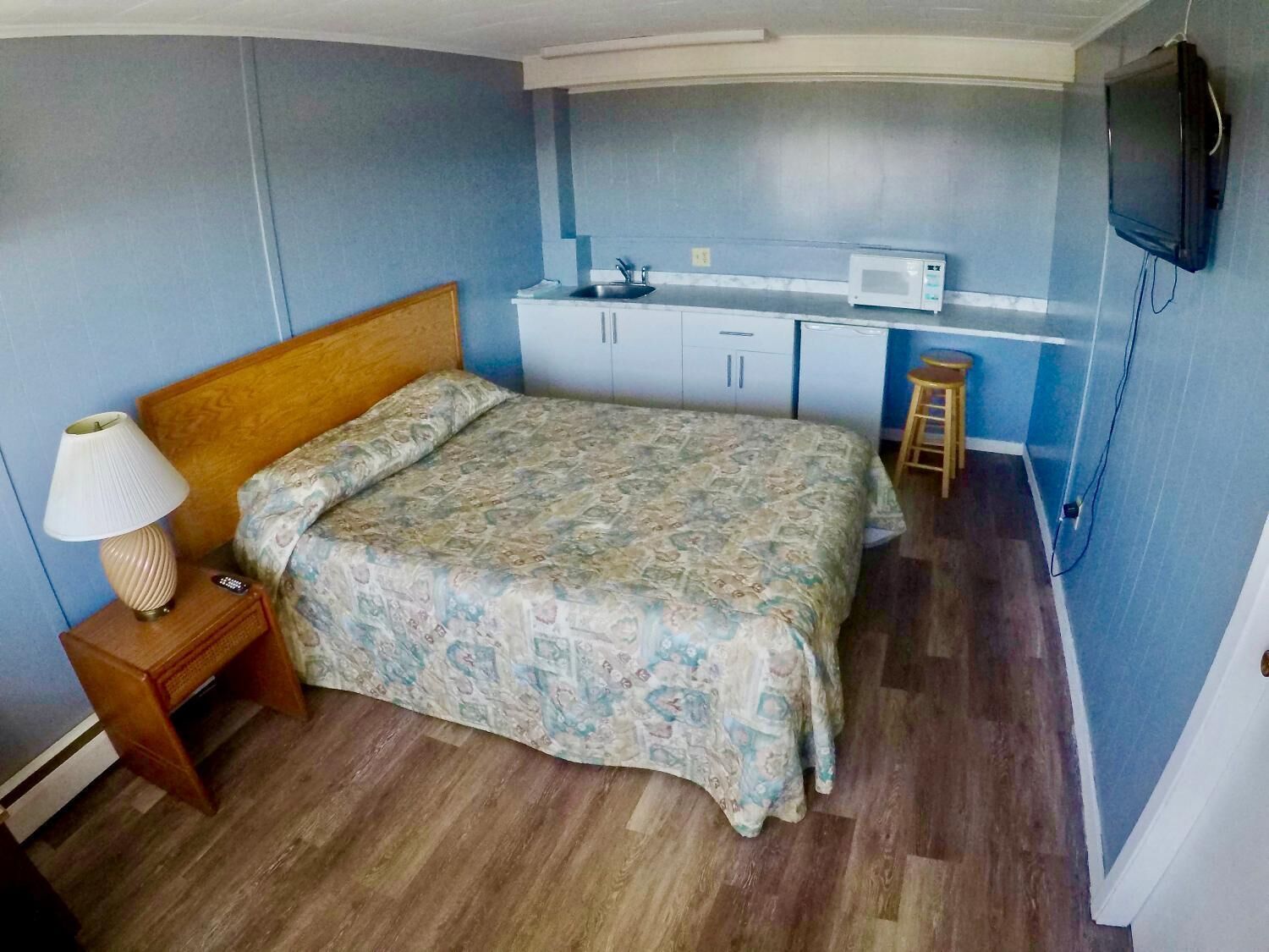A bedroom with a bed, a nightstand, a lamp, a microwave, a small stool, and a TV mounted on the wall.
