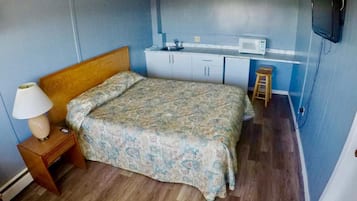 A bedroom with a bed, a nightstand, a lamp, a microwave, a small stool, and a TV mounted on the wall.