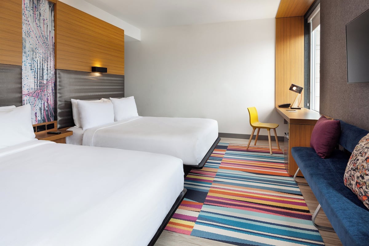 A hotel room with two beds, a desk, a chair, and a colorful striped rug.