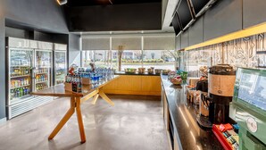 A modern coffee shop interior with a central wooden table, refrigerated beverage display, and a coffee machine.