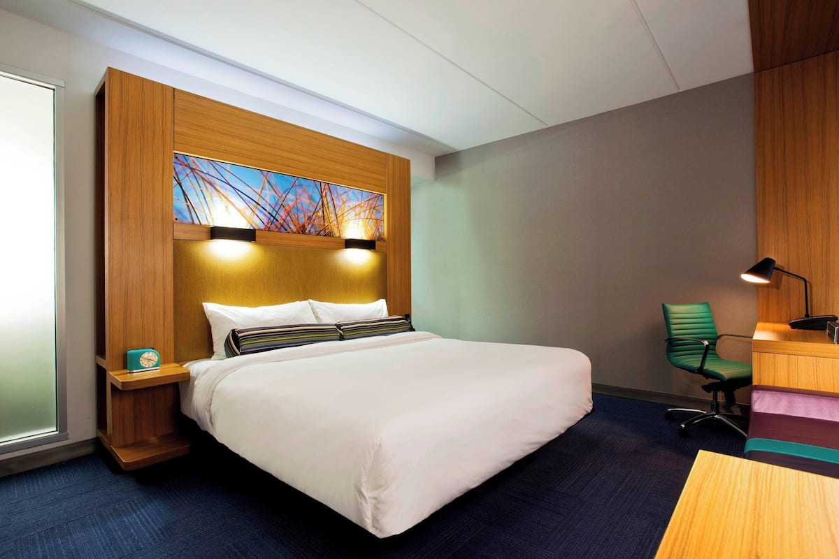 A hotel room with a large bed, a wooden headboard, a nightstand with a clock, a desk with a lamp, and a green chair.