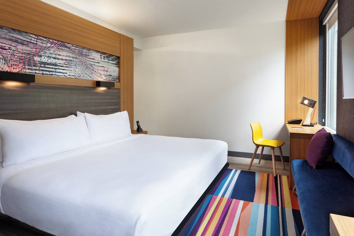 A hotel room with a large bed, a desk with a chair, a colorful striped rug, and a large window with blinds.
