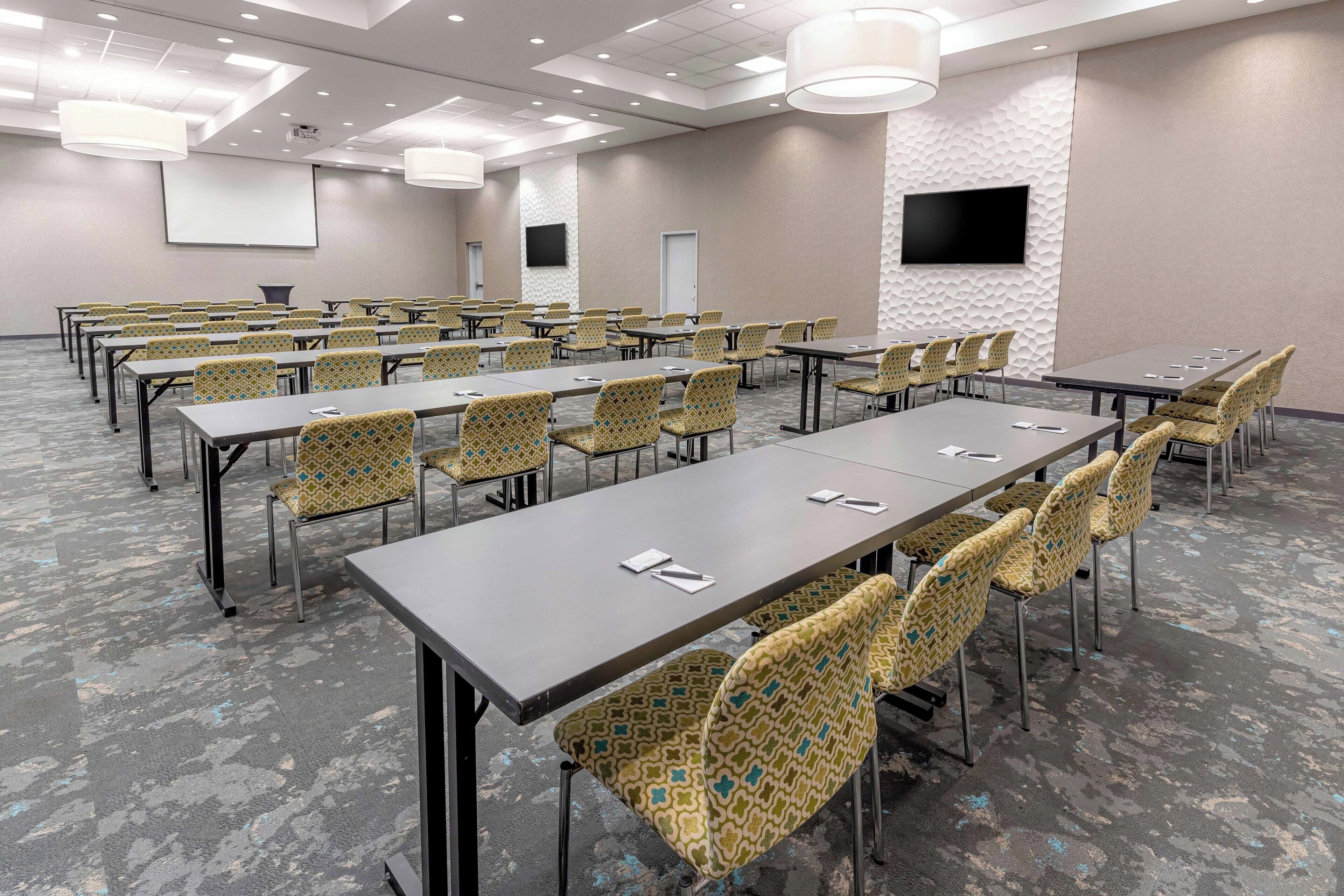 A conference room with multiple tables and chairs, a projector screen, and a flat-screen TV on the wall.