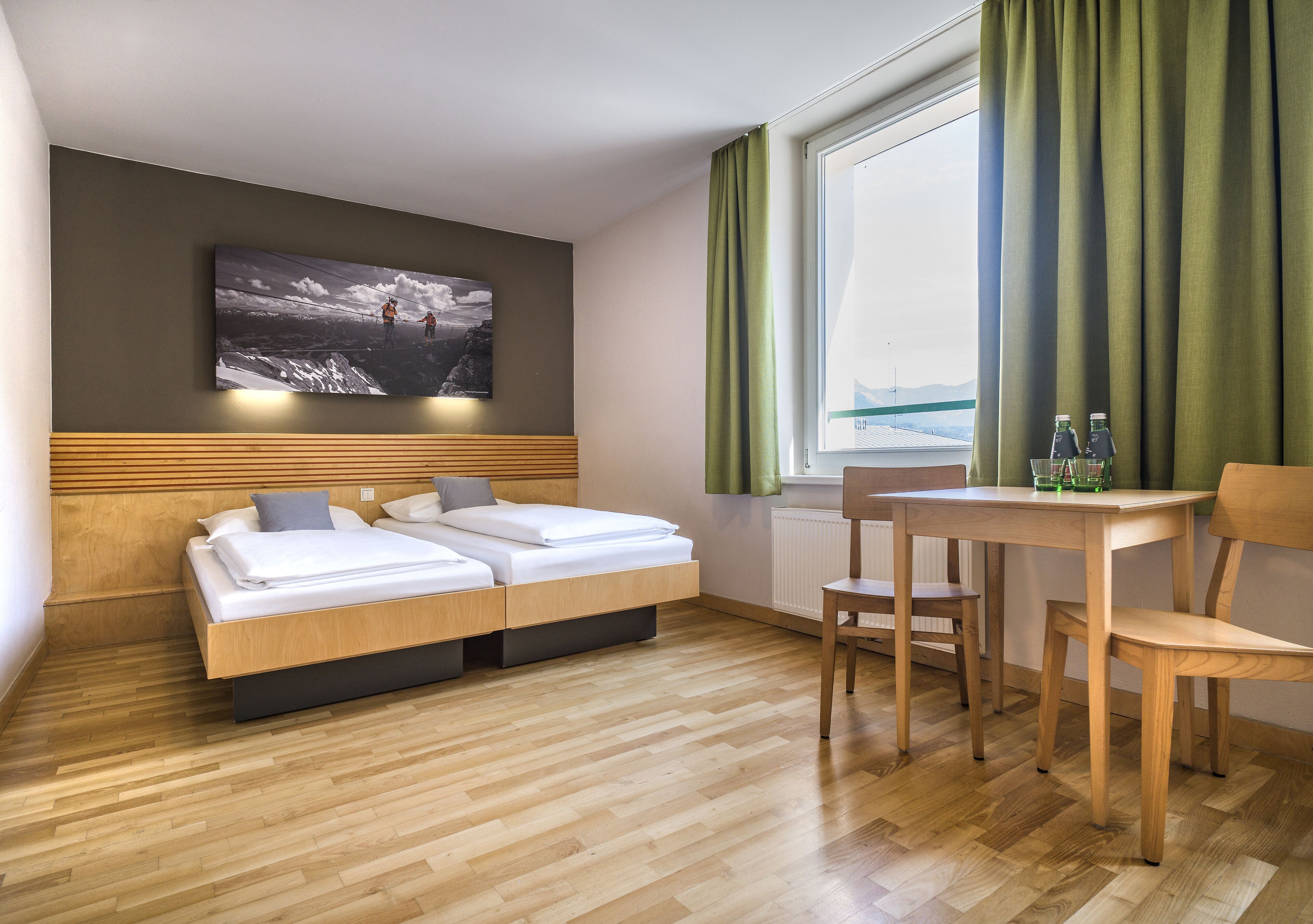 A hotel room with a bed, a small table with chairs, a window with curtains, and a painting on the wall.