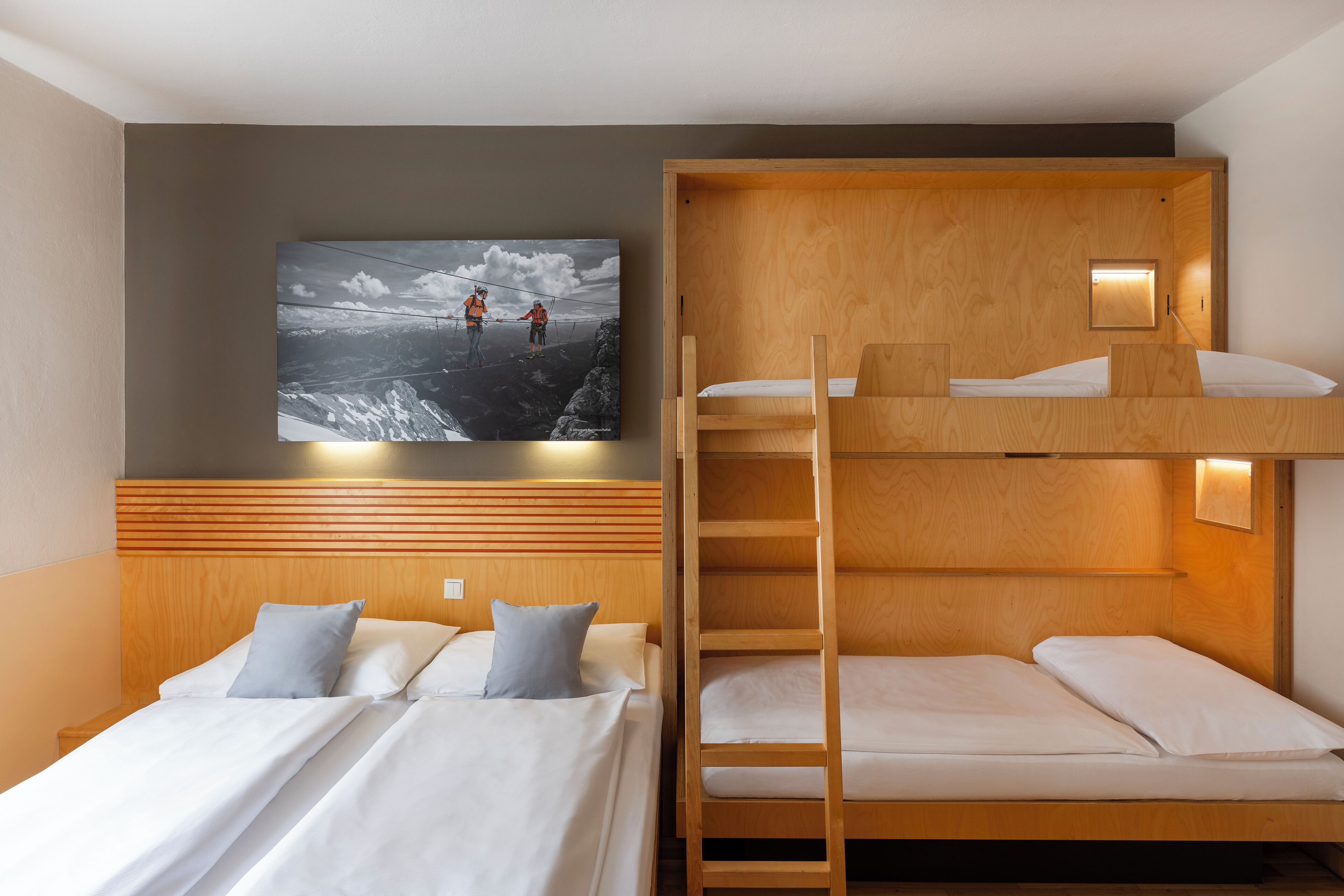 A room with two beds, one with a headboard and the other with a bunk bed. There is a wall-mounted picture of skiers on a mountain.