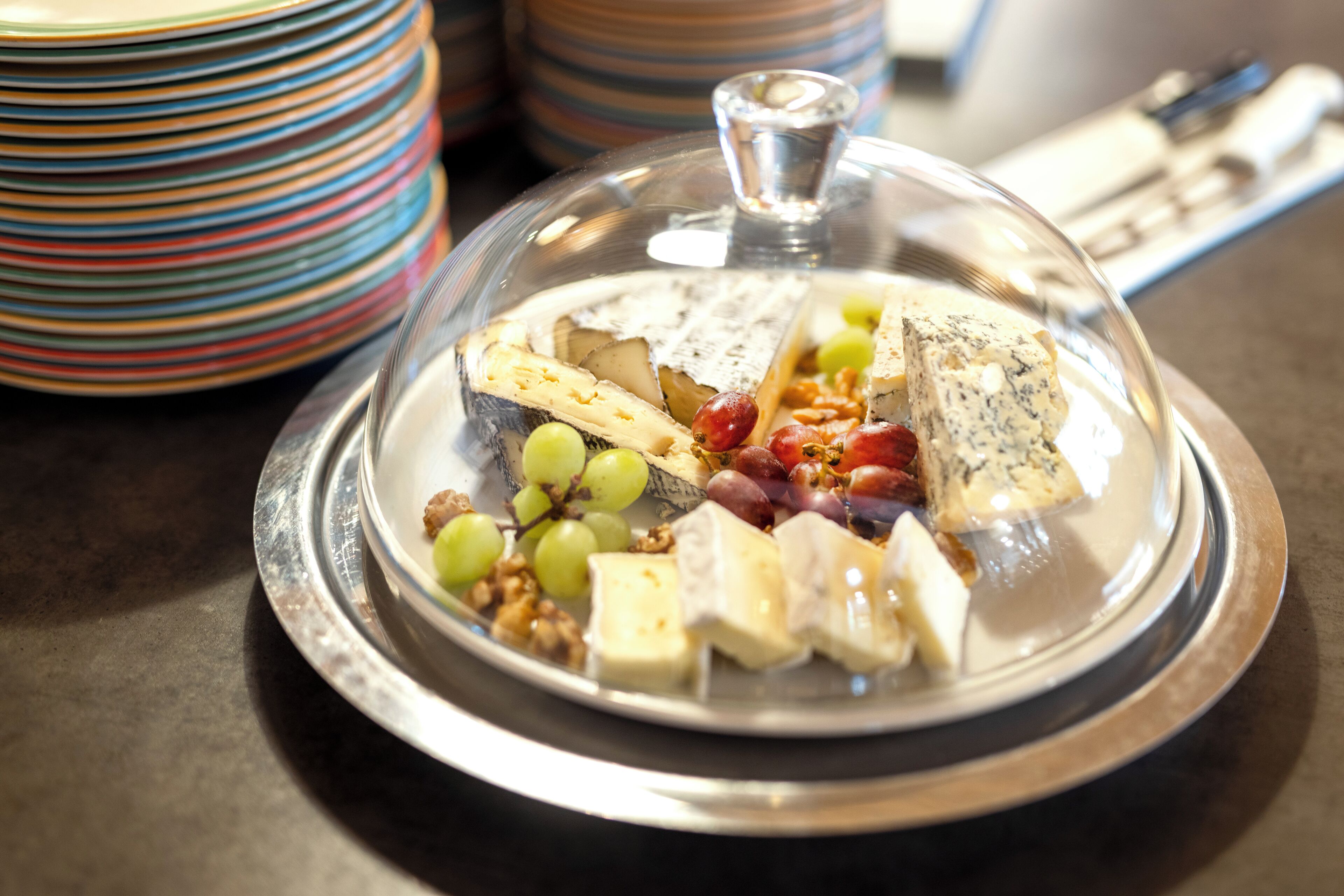 A cheese platter with various cheeses, grapes, and nuts on a silver platter with a glass dome cover.