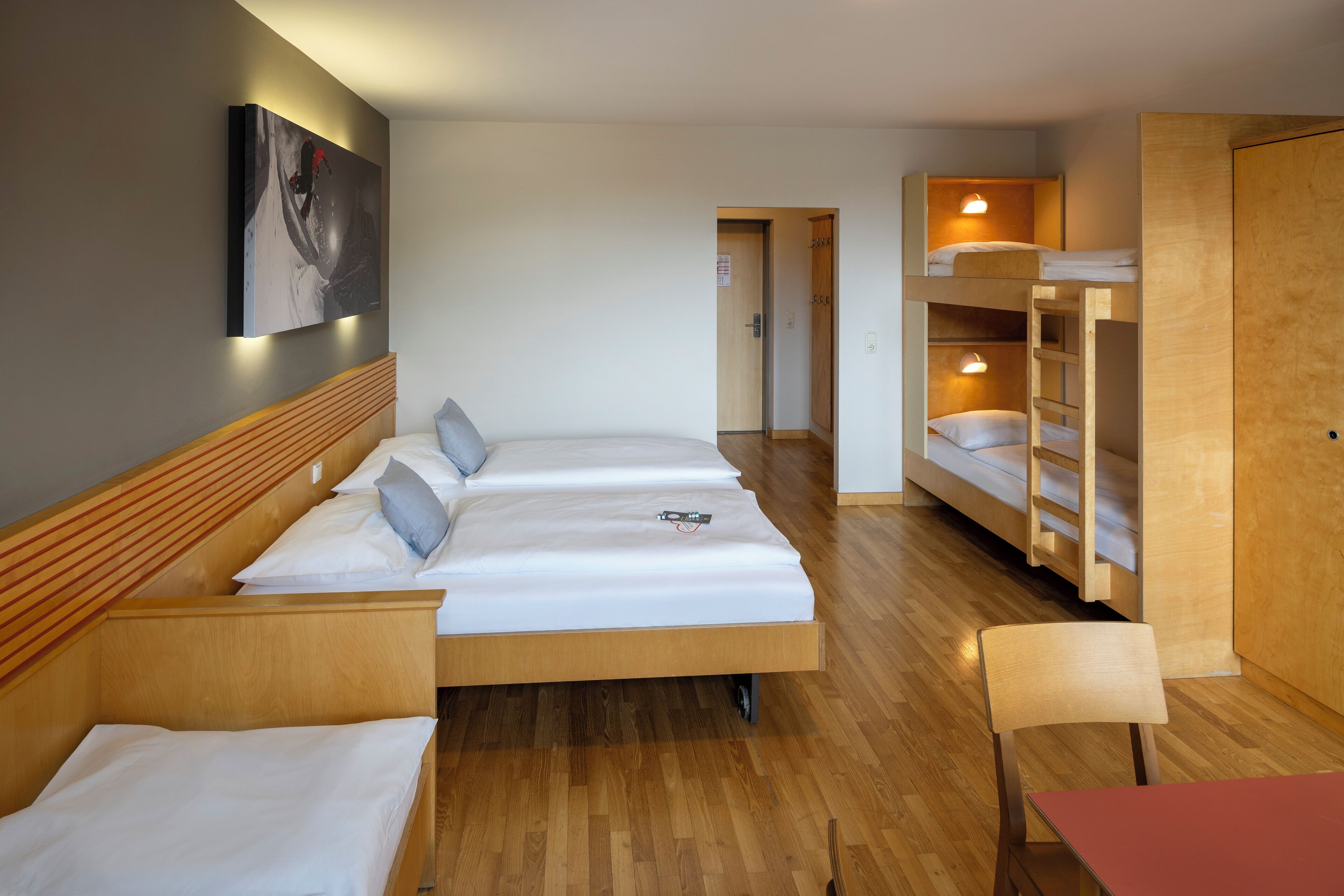 A hotel room with three beds, a wooden headboard, a red table, and a bunk bed with lights.