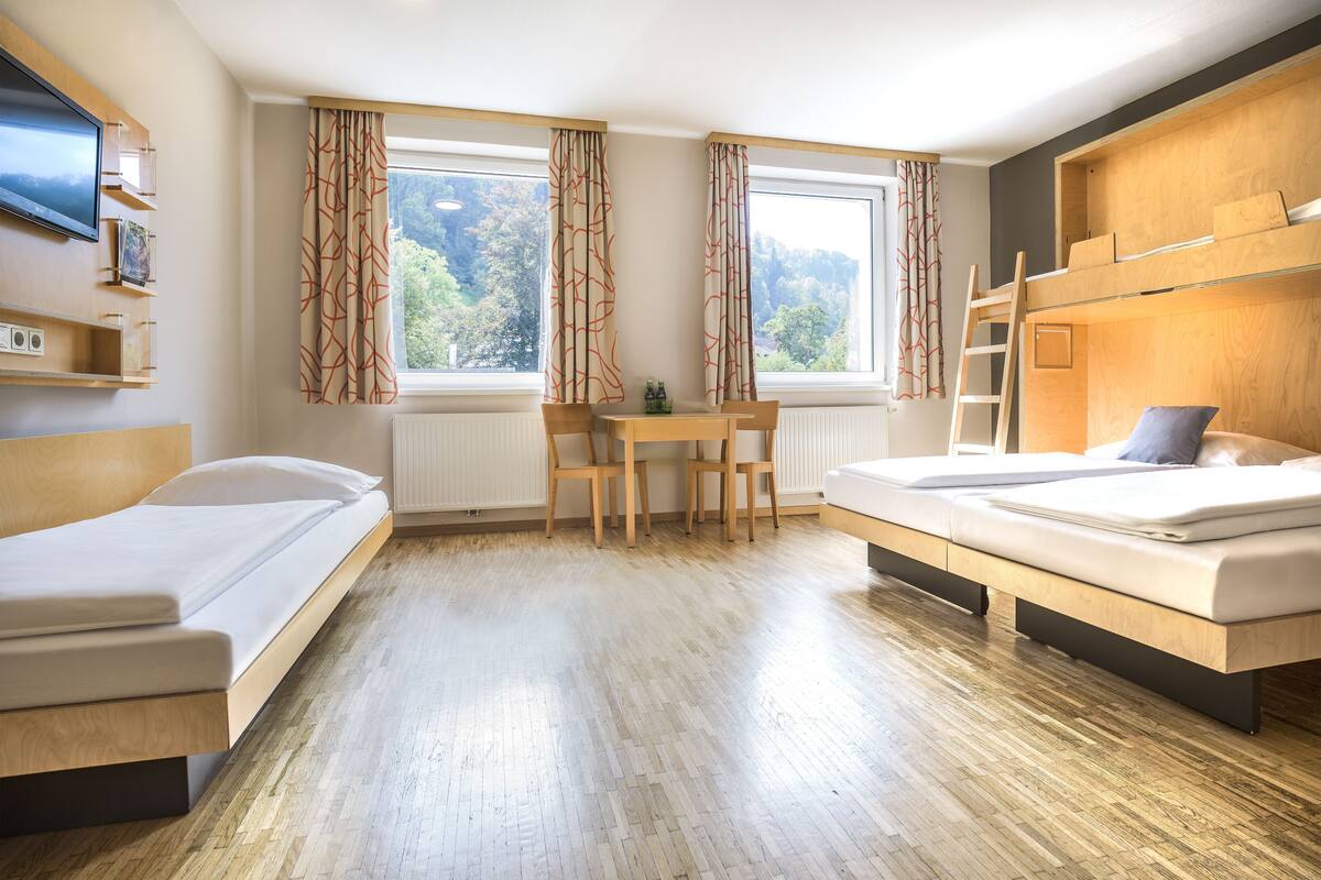 Una habitació d'hotel moderna amb sòl de fusta, una taula de treball amb cadires, un llit superposat i un televisor penjat a la paret.