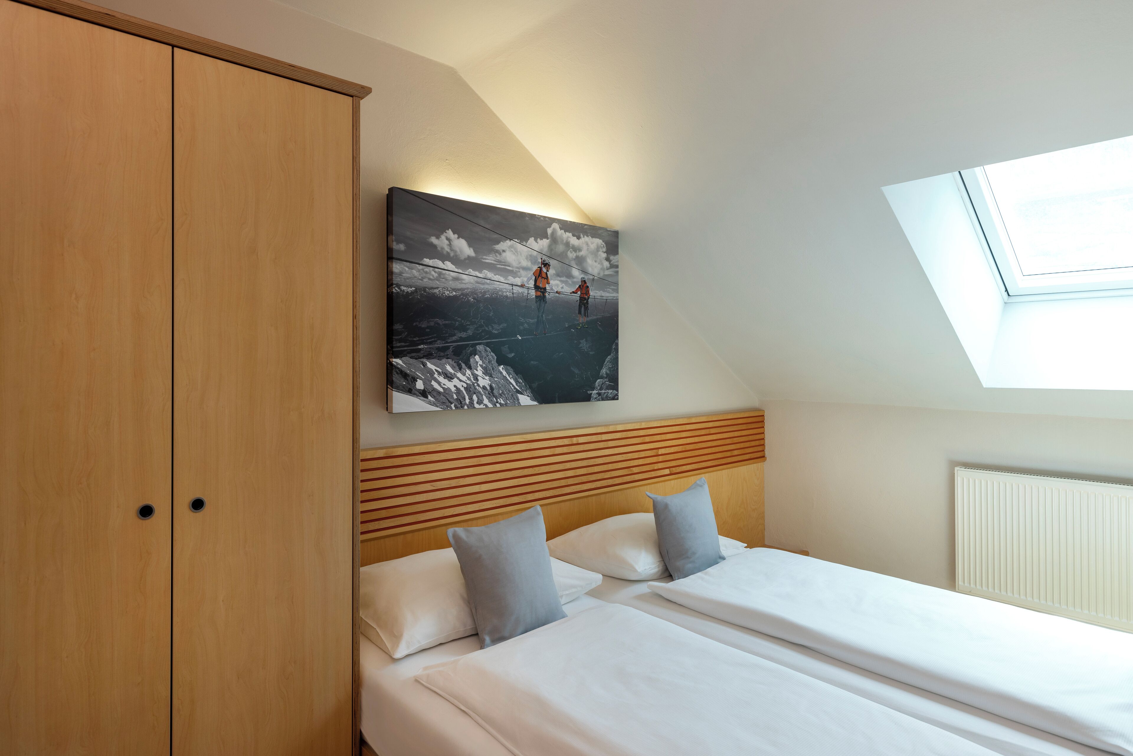 Une chambre avec une armoire en bois, un lit avec du linge de lit blanc, une tête de lit avec des lattes de bois horizontales, un puits de lumière et un tableau accroché au mur représentant deux personnes sur un pont.