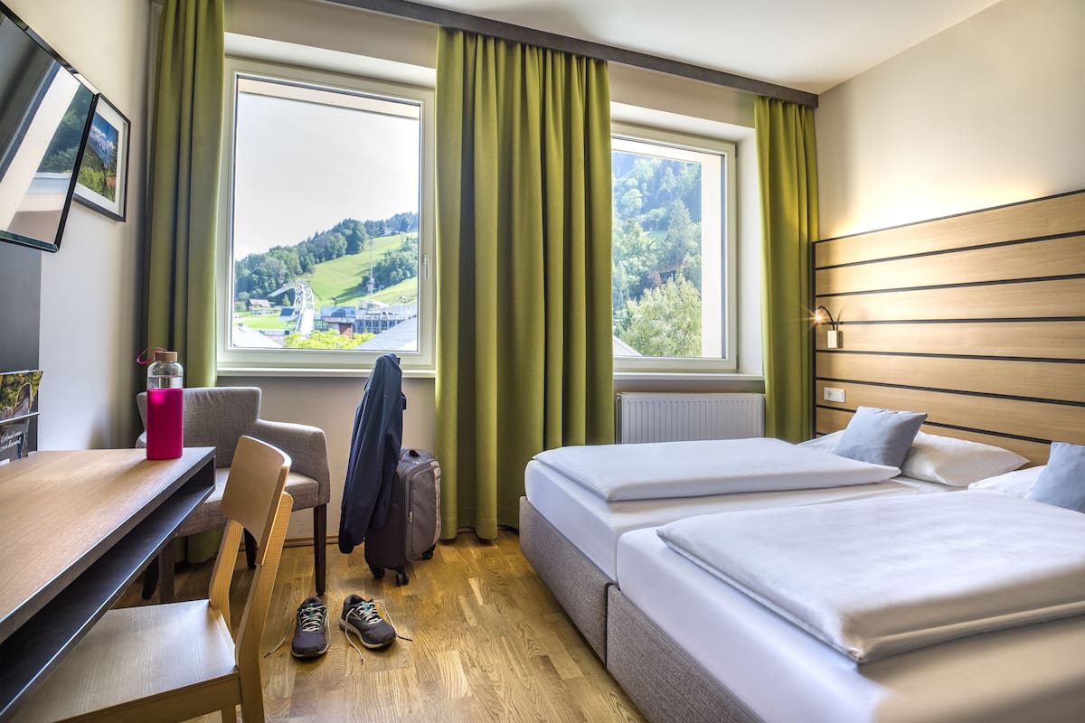 Habitació d'hotel amb dos llits, un escriptori, una cadira, una maleta i vista a un paisatge verd a través de la finestra.