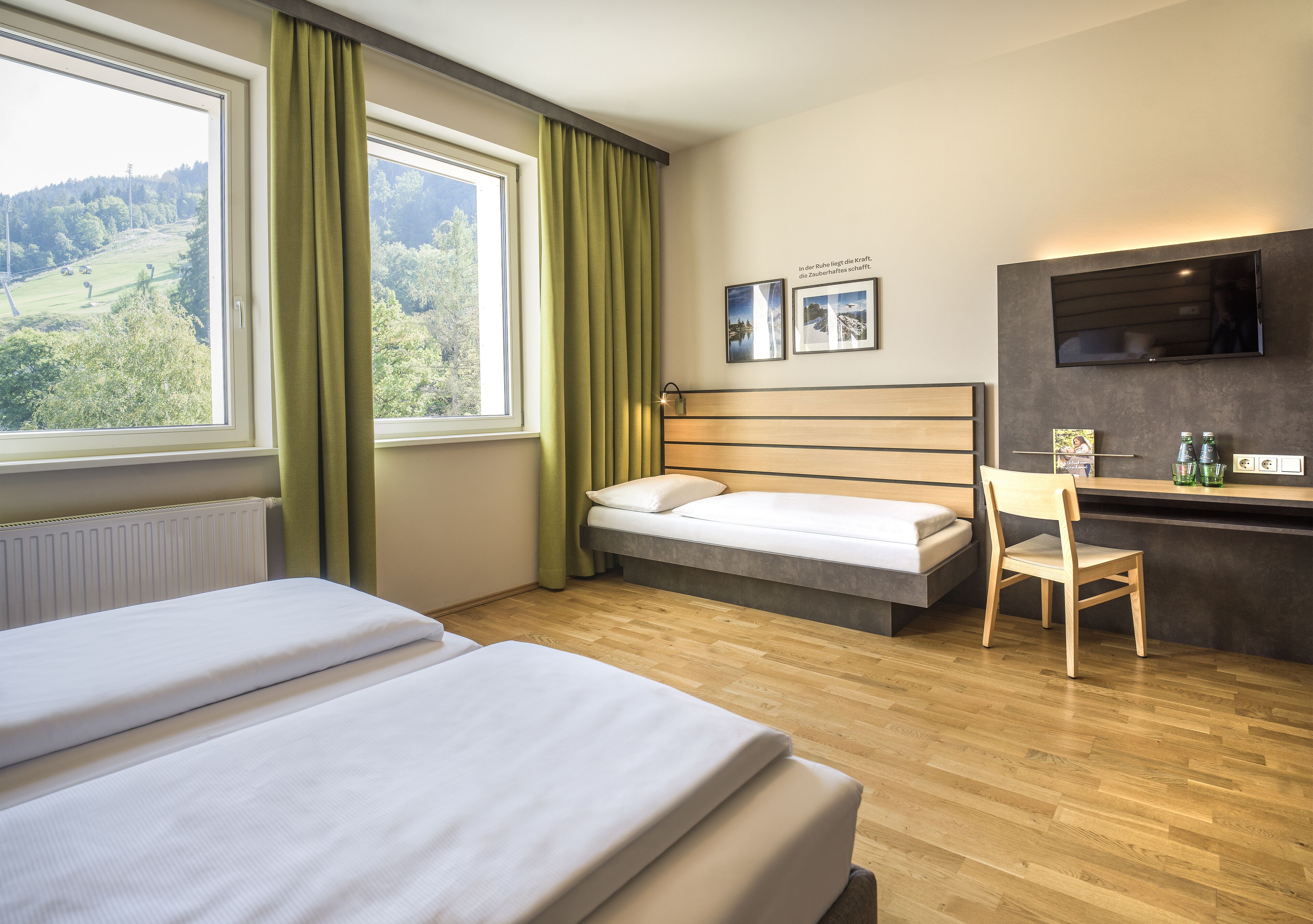A hotel room with two beds, a desk, a chair, a TV, and a view of a green landscape.