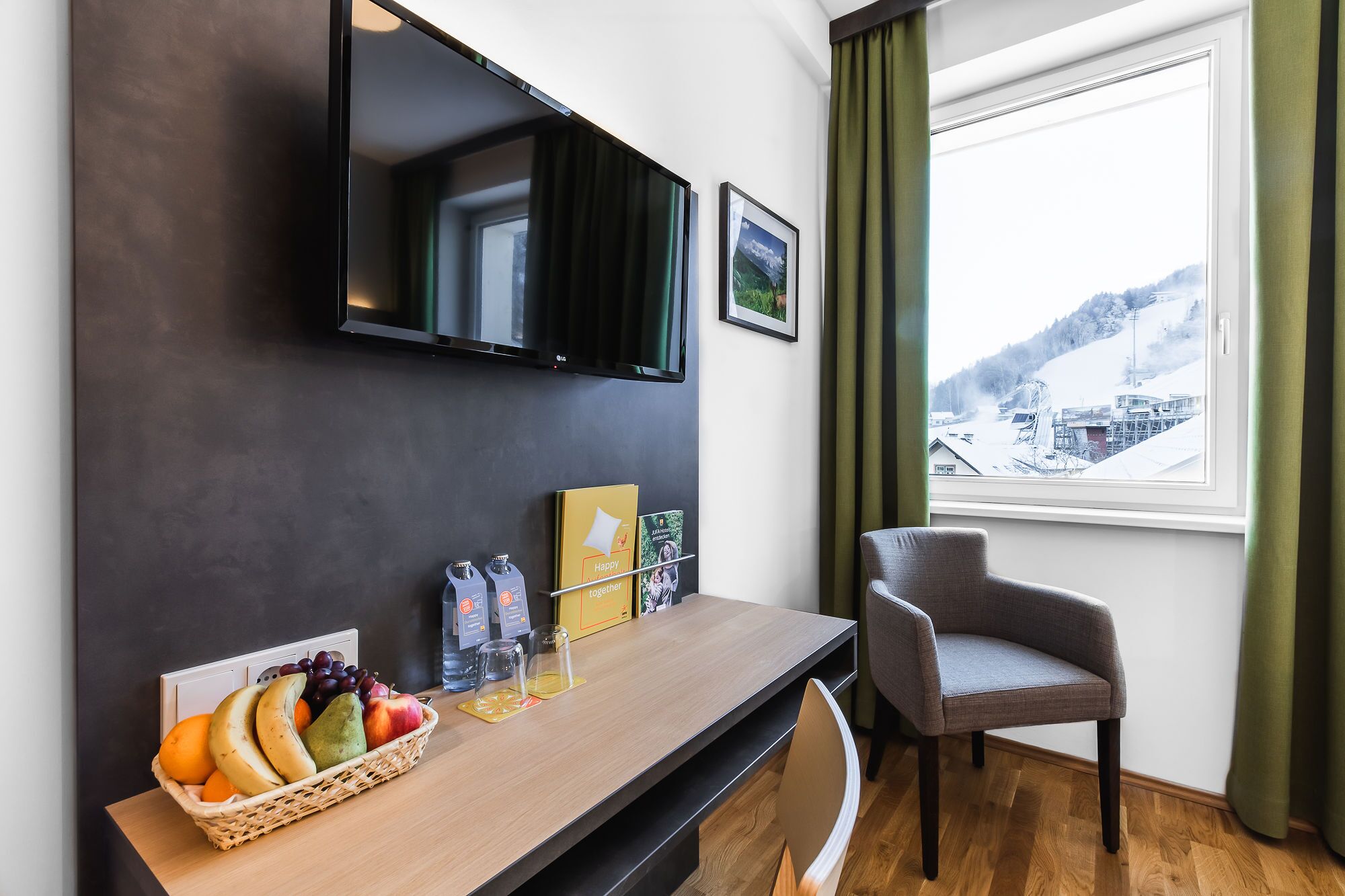 A hotel room with a wooden desk, a television mounted on the wall, a chair, and a window with a view of a snowy landscape.
