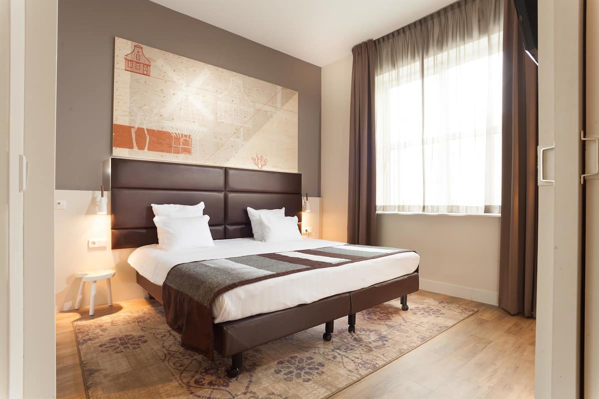 Quarto de hotel moderno com uma cama grande, cabeceira com design de paisagem urbana, um banquinho pequeno e uma janela ampla com cortinas.