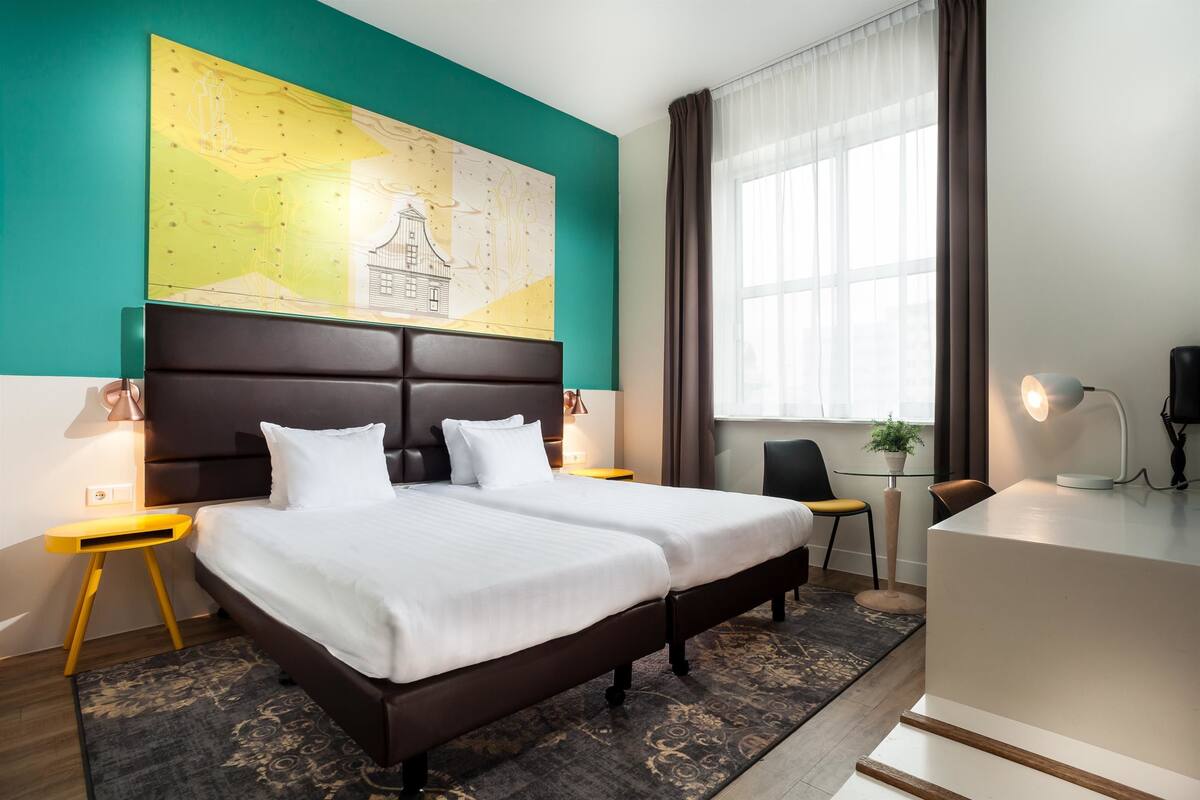 Quarto de hotel com uma cama grande, uma escrivaninha, uma cadeira, um mapa na parede e uma janela com cortinas.