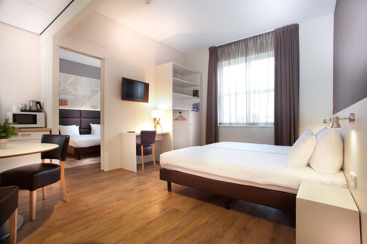 Quarto de hotel com cama, escrivaninha, cadeira, televisão e janela com cortinas.