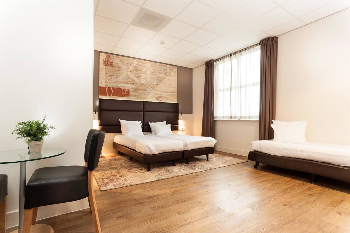 Quarto de hotel moderno com uma cama grande, uma cama menor, uma mesa de vidro, uma cadeira e um vaso de planta.