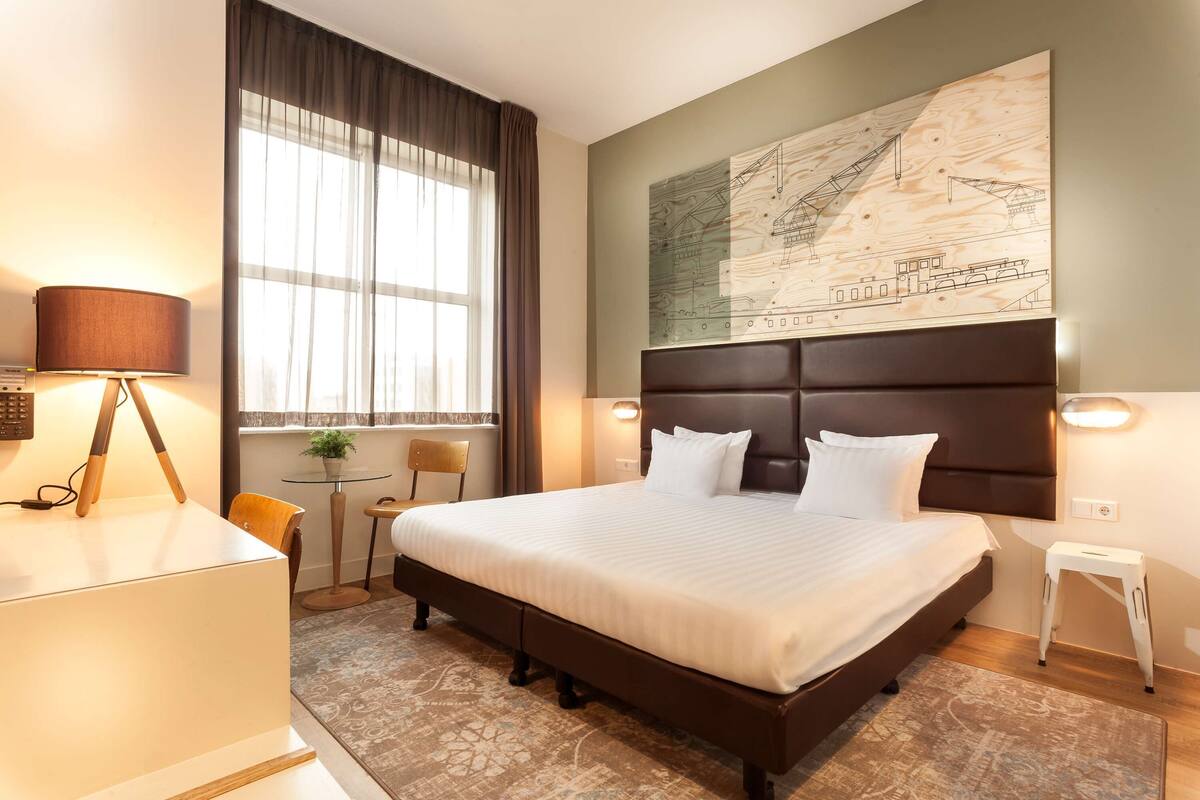 Quarto de hotel com uma cama grande, uma escrivaninha com luminária, uma mesinha com uma planta e uma grande obra de arte com mapa na parede.