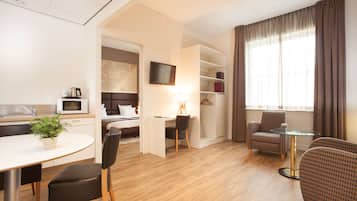 Een moderne hotelkamer met een houten vloer, een zithoek met een tafel en stoelen, een werkplek met een bureau en stoel, een bed met hoofdeinde, een aan de muur bevestigde televisie en een groot raam met gordijnen.