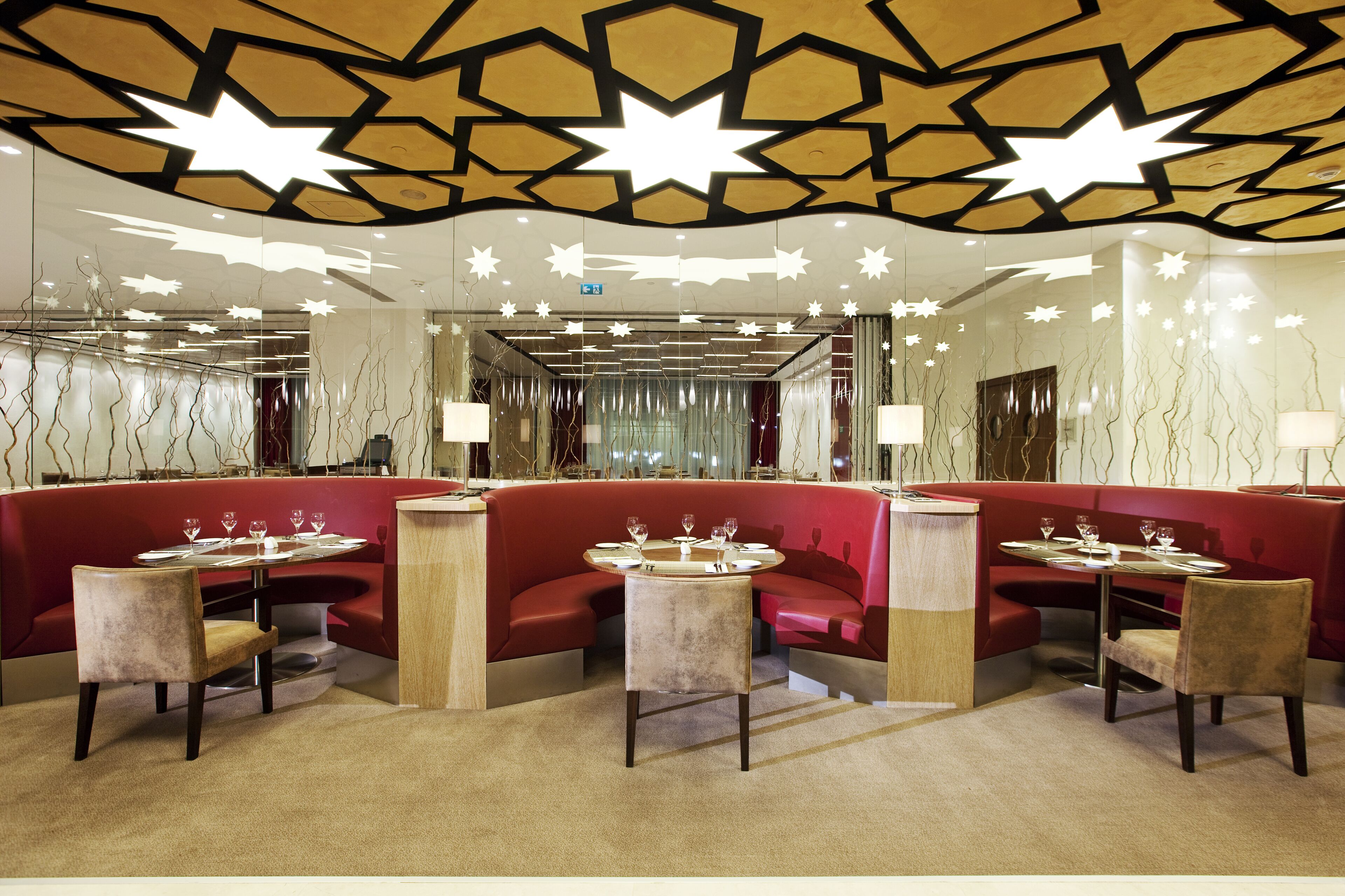 Un restaurante moderno con puestos tapizados en rojo y mesas redondas, que presenta un diseño geométrico en el techo y superficies reflectantes.