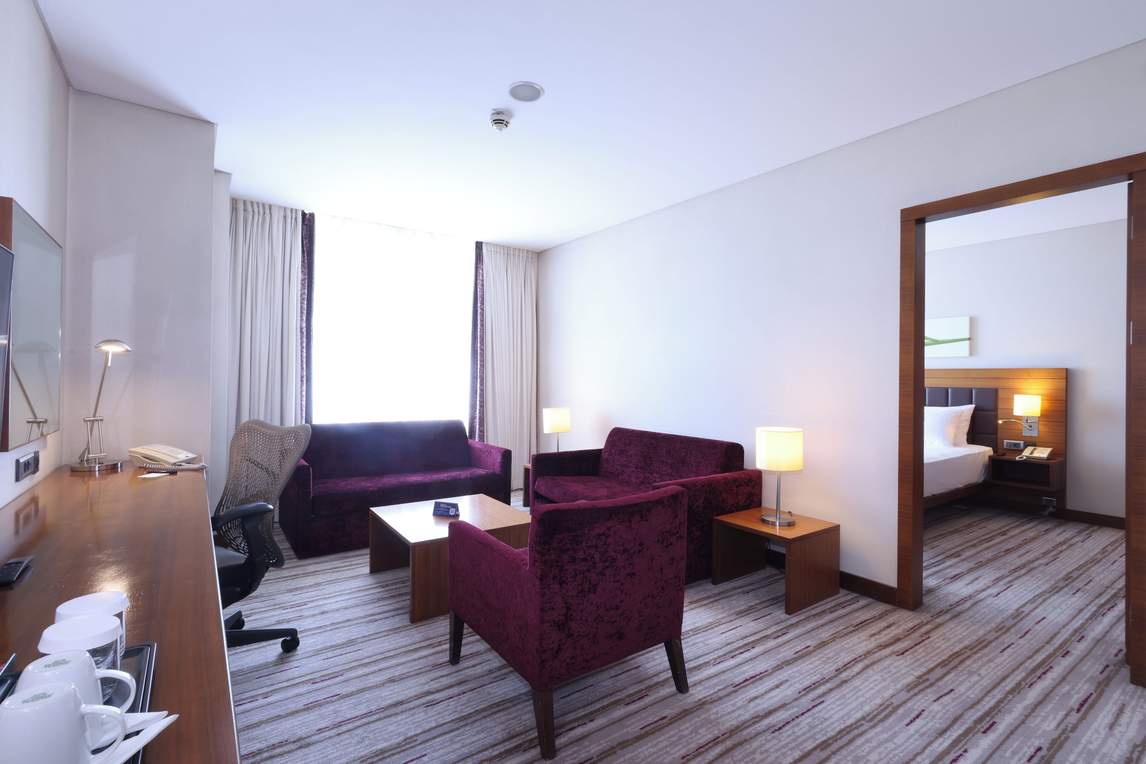 A hotel room with a wooden desk, purple armchairs, a sofa, a bed with a nightstand, and a large mirror.