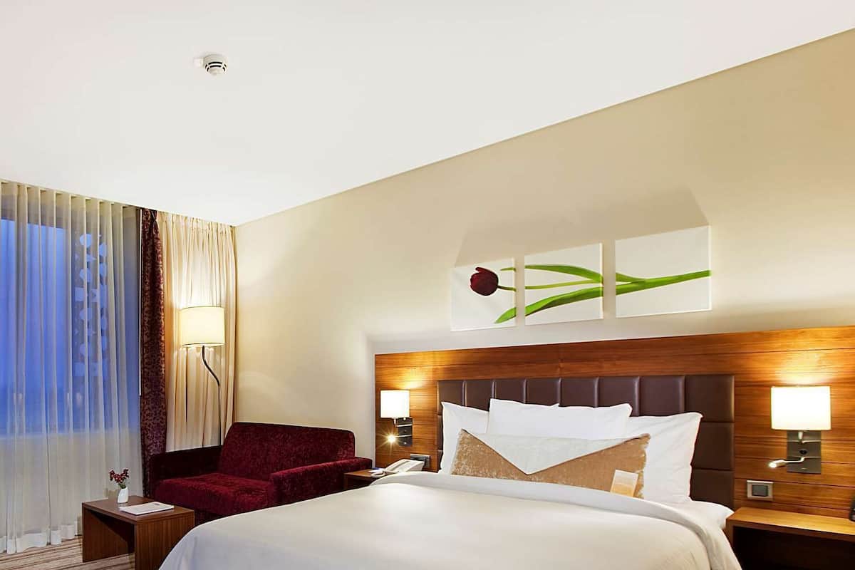 A hotel room with a large bed, a red armchair, a nightstand, and a wall-mounted lamp.