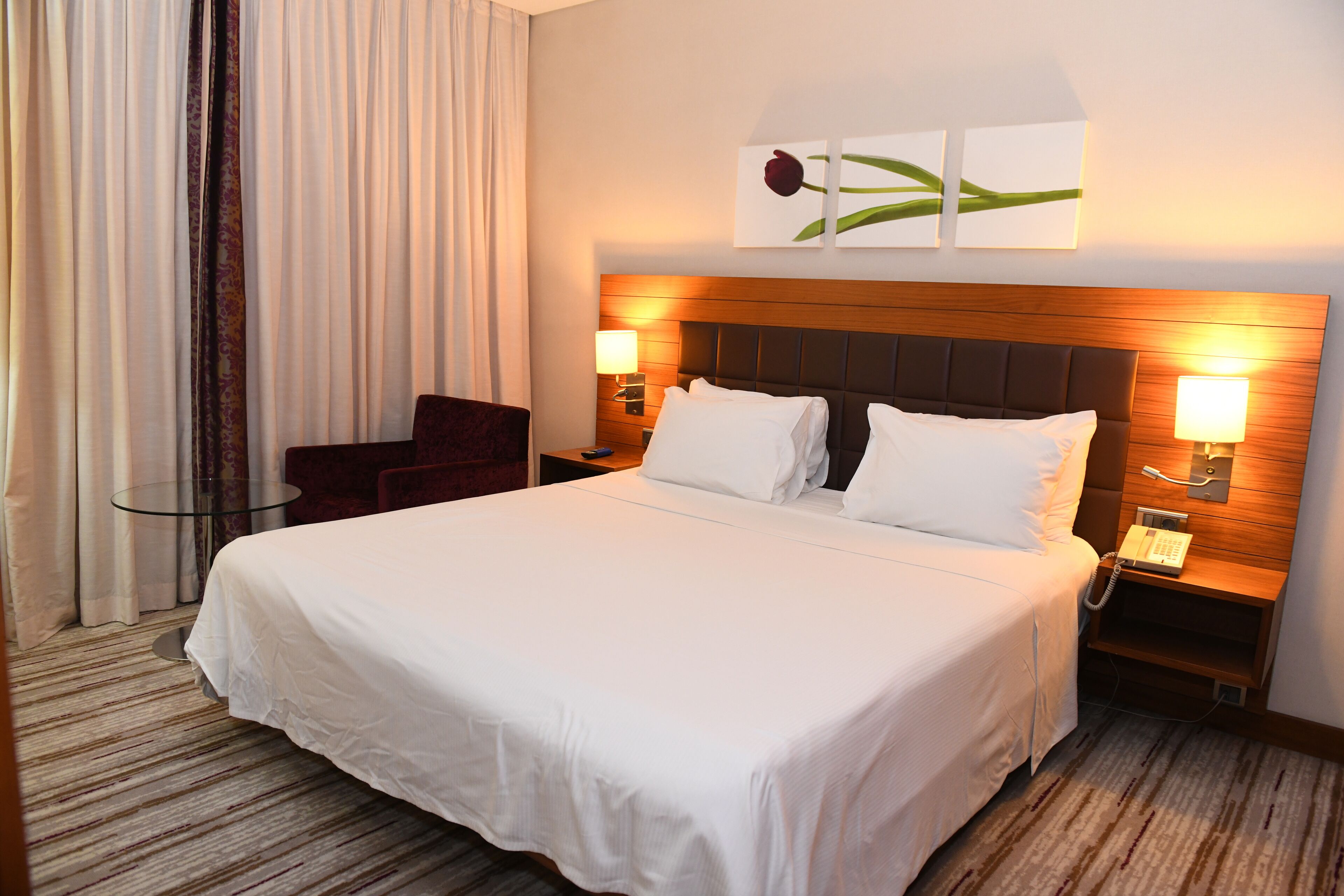 A hotel room with a large bed, two bedside lamps, a telephone, a glass table, a chair, and a curtain.
