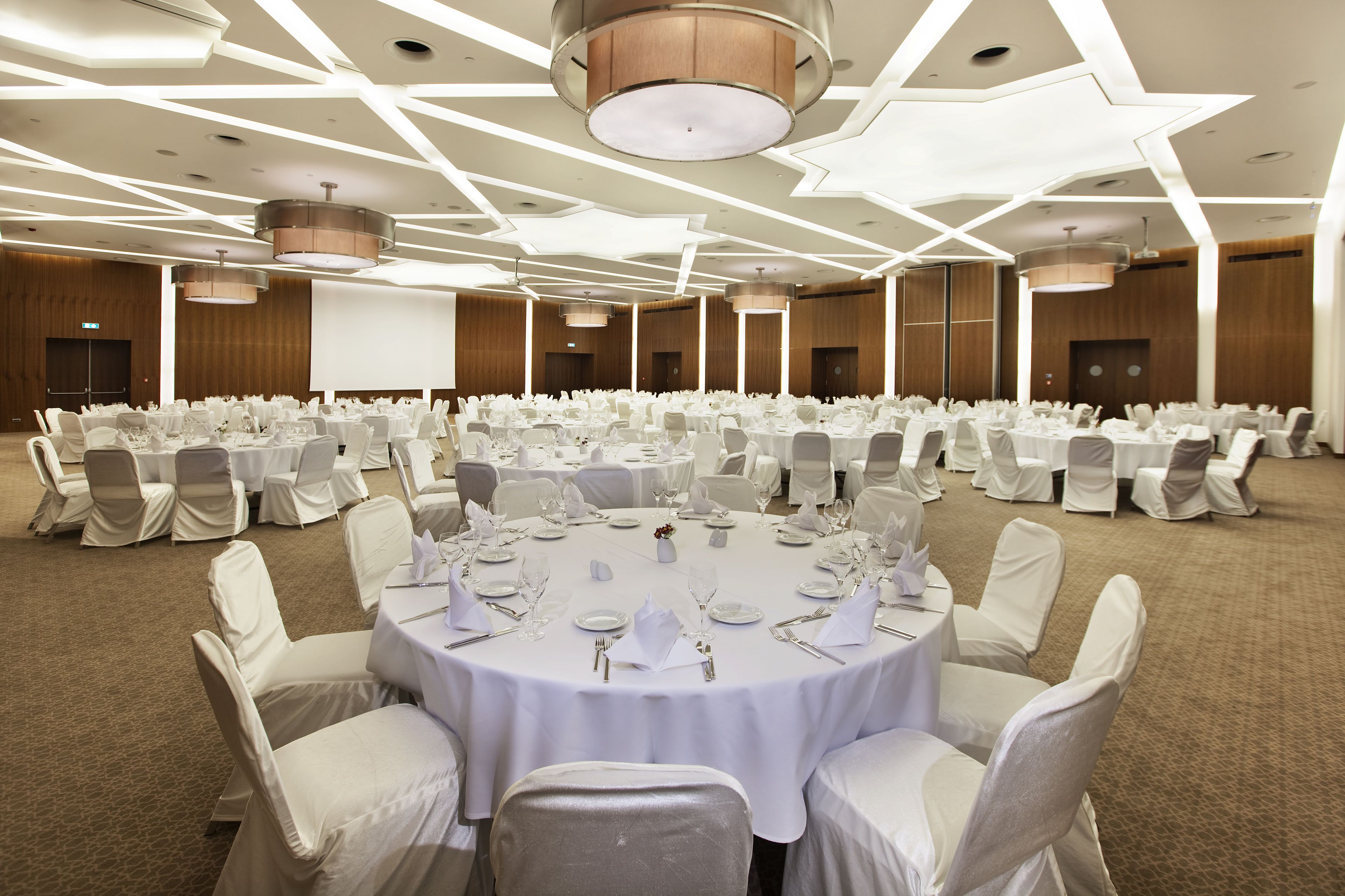 Un amplio salón de conferencias con mesas redondas dispuestas para un evento formal, con un diseño moderno del techo y gran capacidad de asientos.