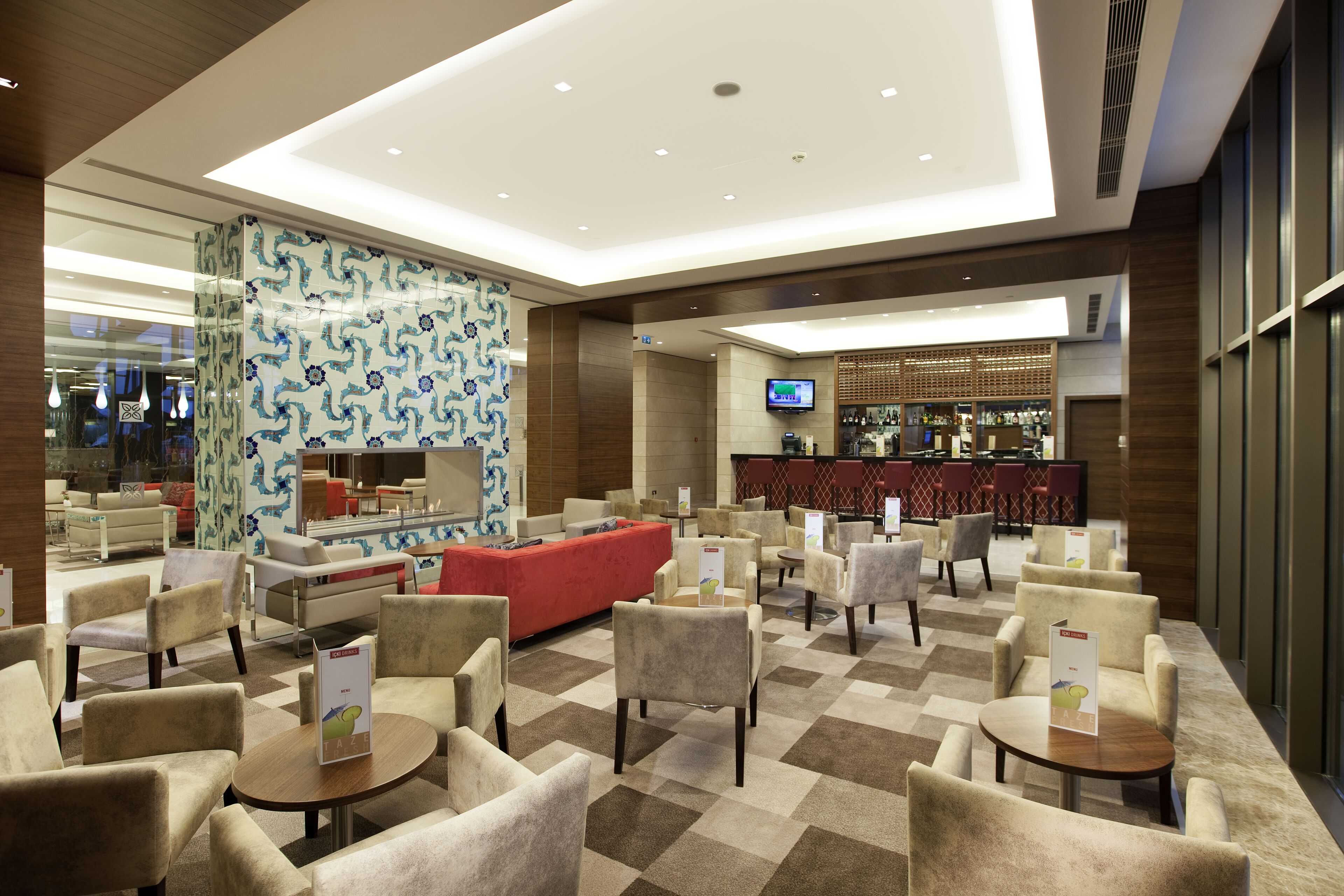 Un vestíbulo de hotel con suelo a cuadros, varias zonas de estar, un sofá rojo y un área de bar con televisor.