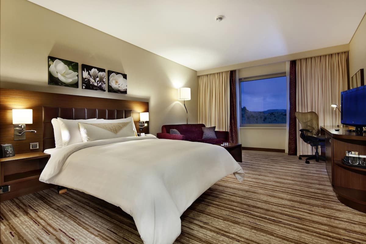 A hotel room with a large bed, a red sofa, a desk with a computer, and a window with curtains.