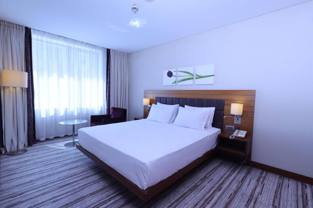 A hotel room with a large bed, a bedside table with a lamp, a small table, and a wall-mounted phone.