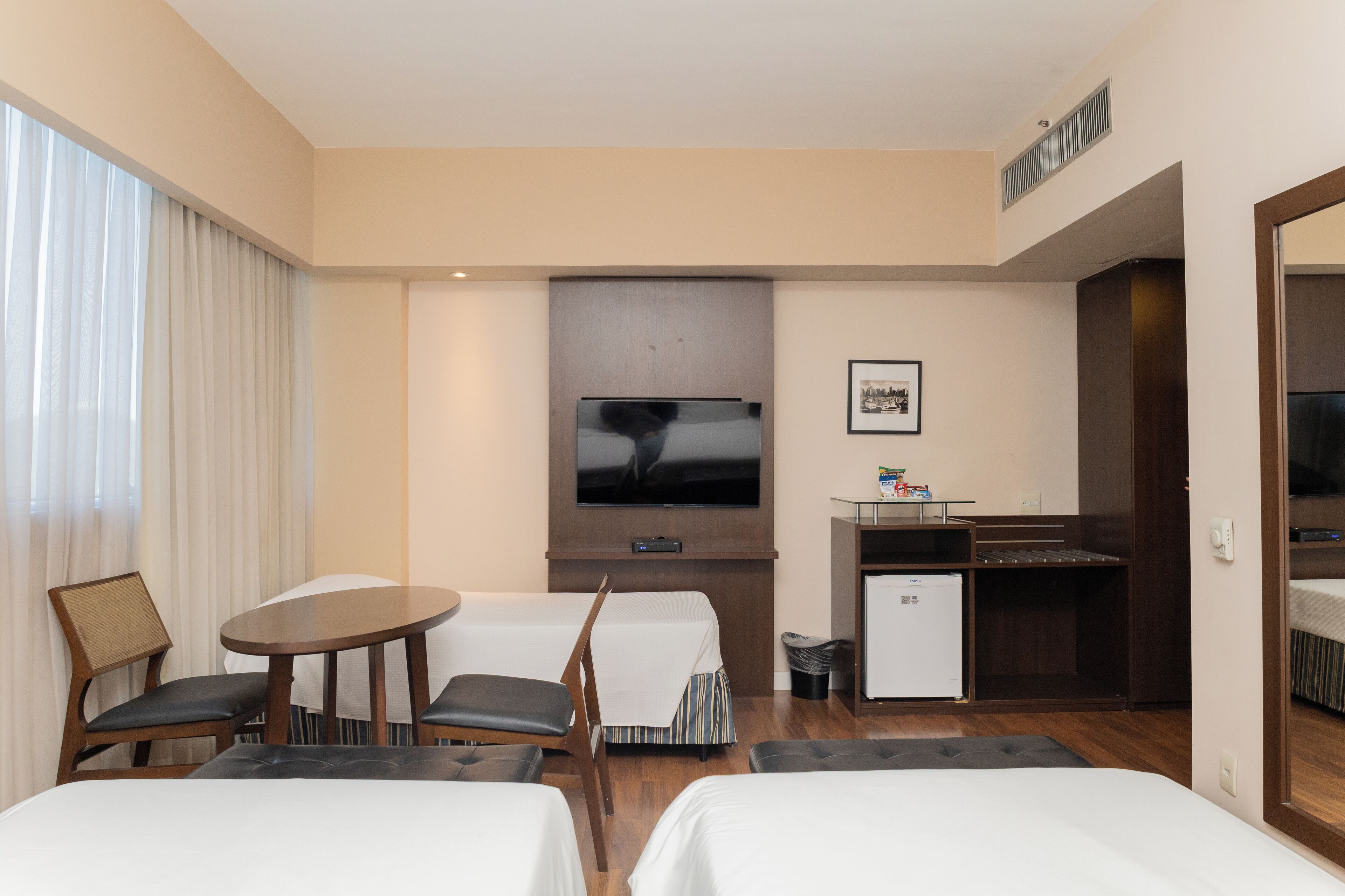 Quarto de hotel com duas camas, uma secretária pequena, televisão e janela com cortinas.