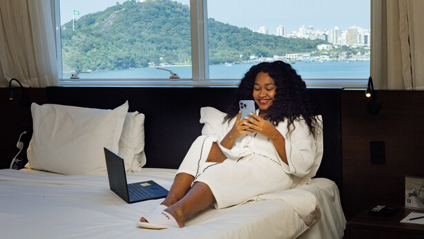 A person sitting on a bed with a laptop, holding a smartphone, in a room with a large window overlooking a city and a hill. - Golden Tulip Porto Vitória (Vitoria)