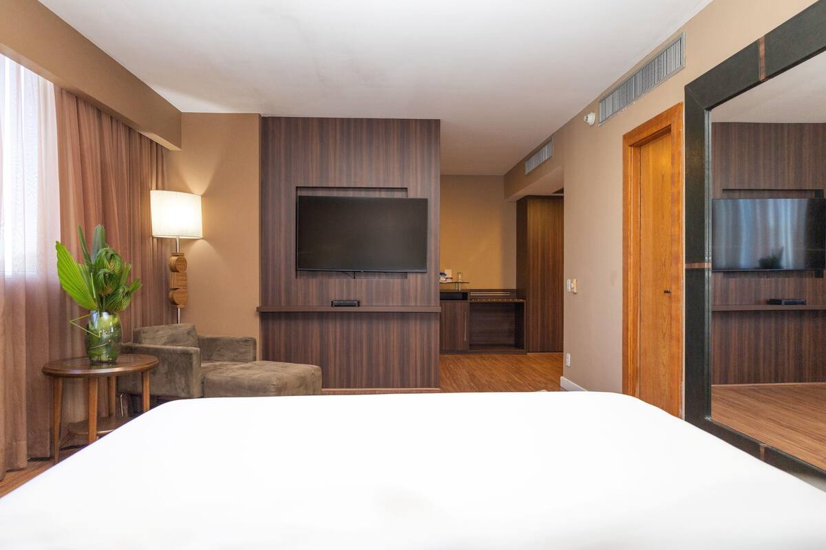 A hotel room with a large bed, a flat-screen TV mounted on the wall, a small round table with a plant, a sofa, and a wooden door.