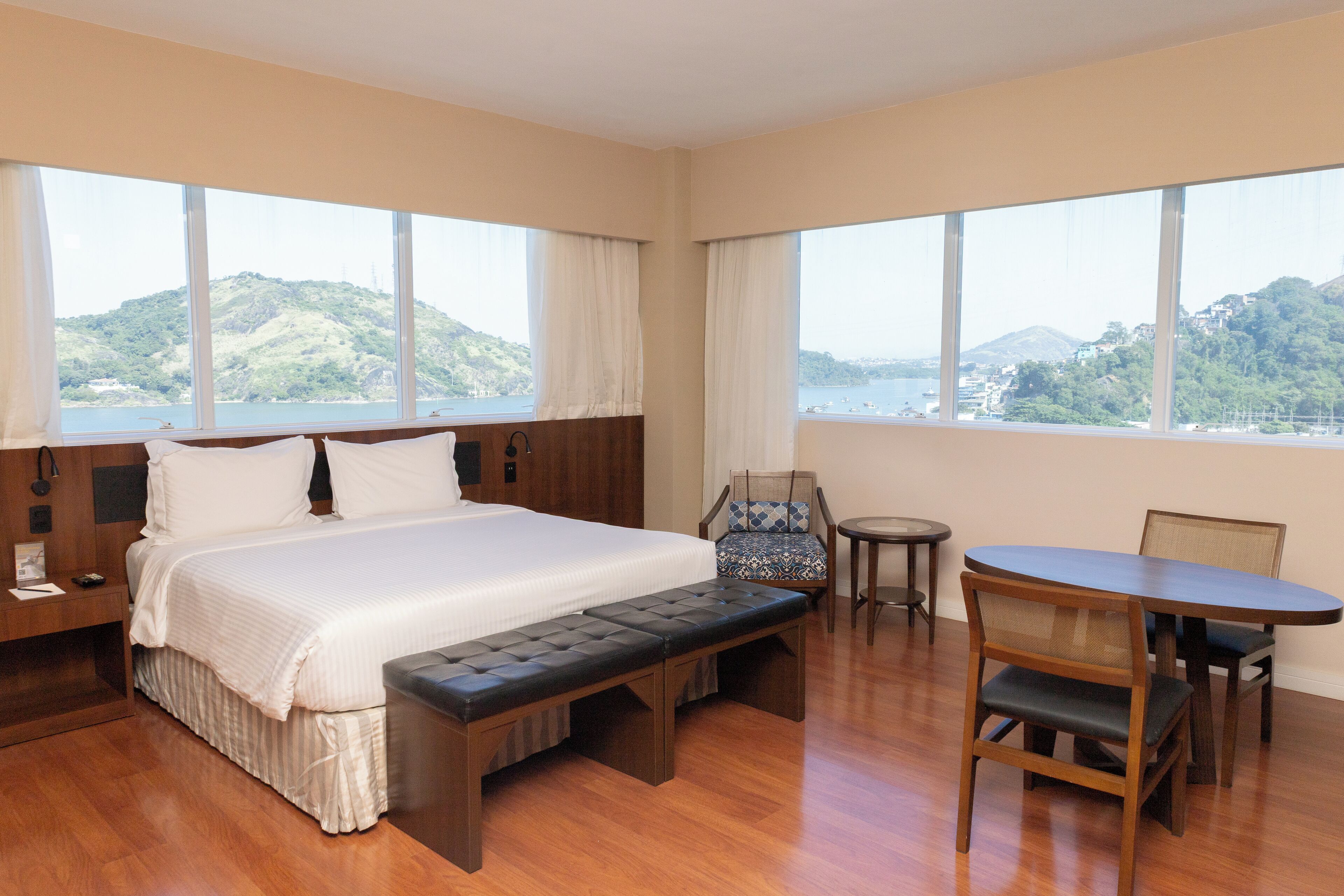 Quarto de hotel com uma cama grande, um banco, uma mesa de jantar e cadeiras. Janelas amplas oferecem vista para colinas e um corpo de água.